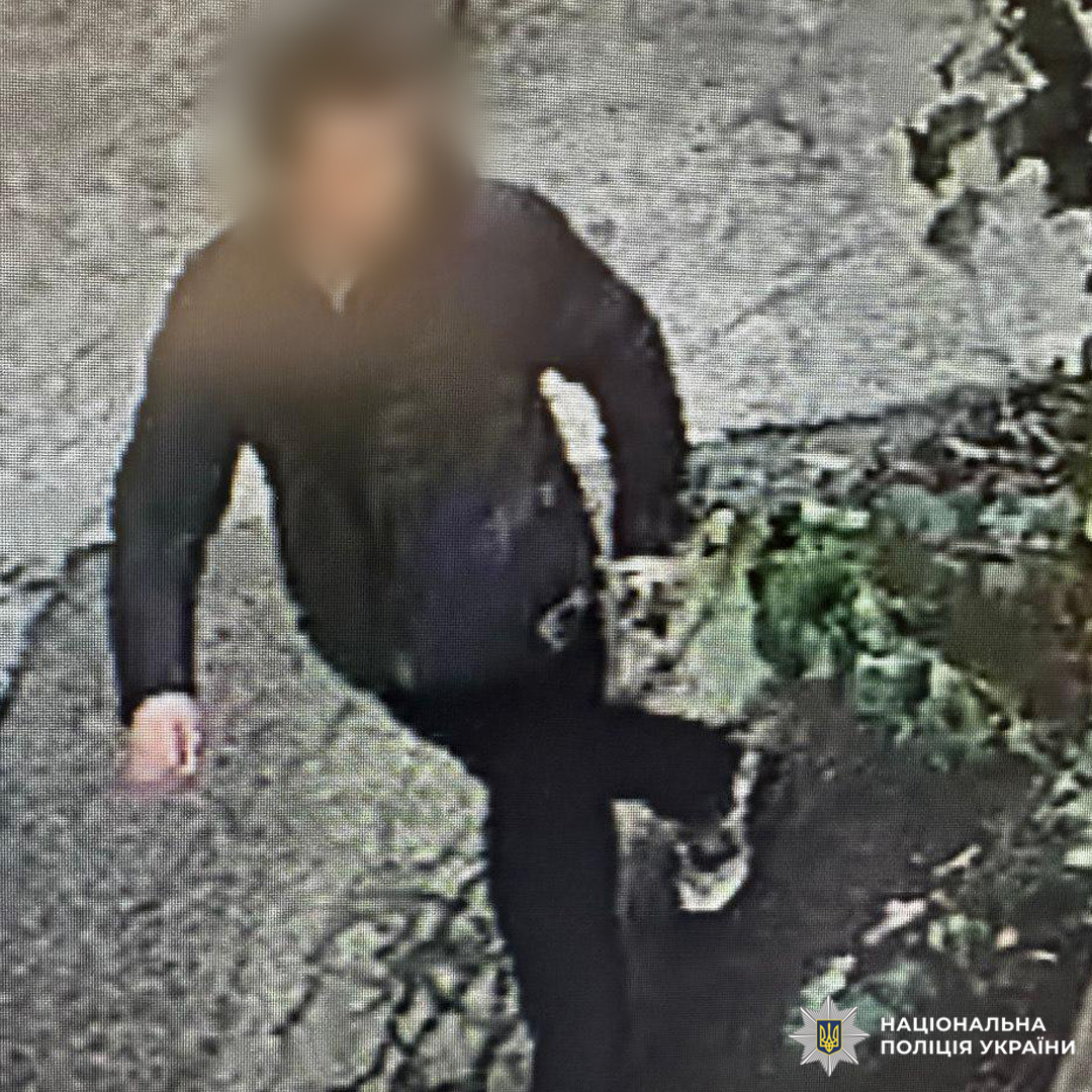 Чоловік у темному одязі, зафіксований камерою спостереження під час руху по асфальтованій ділянці поблизу зелених насаджень.