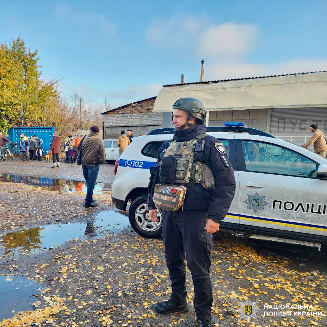 Поліцейський у бронежилеті біля службового авто, група людей біля синьої споруди, осінь, мокра земля.