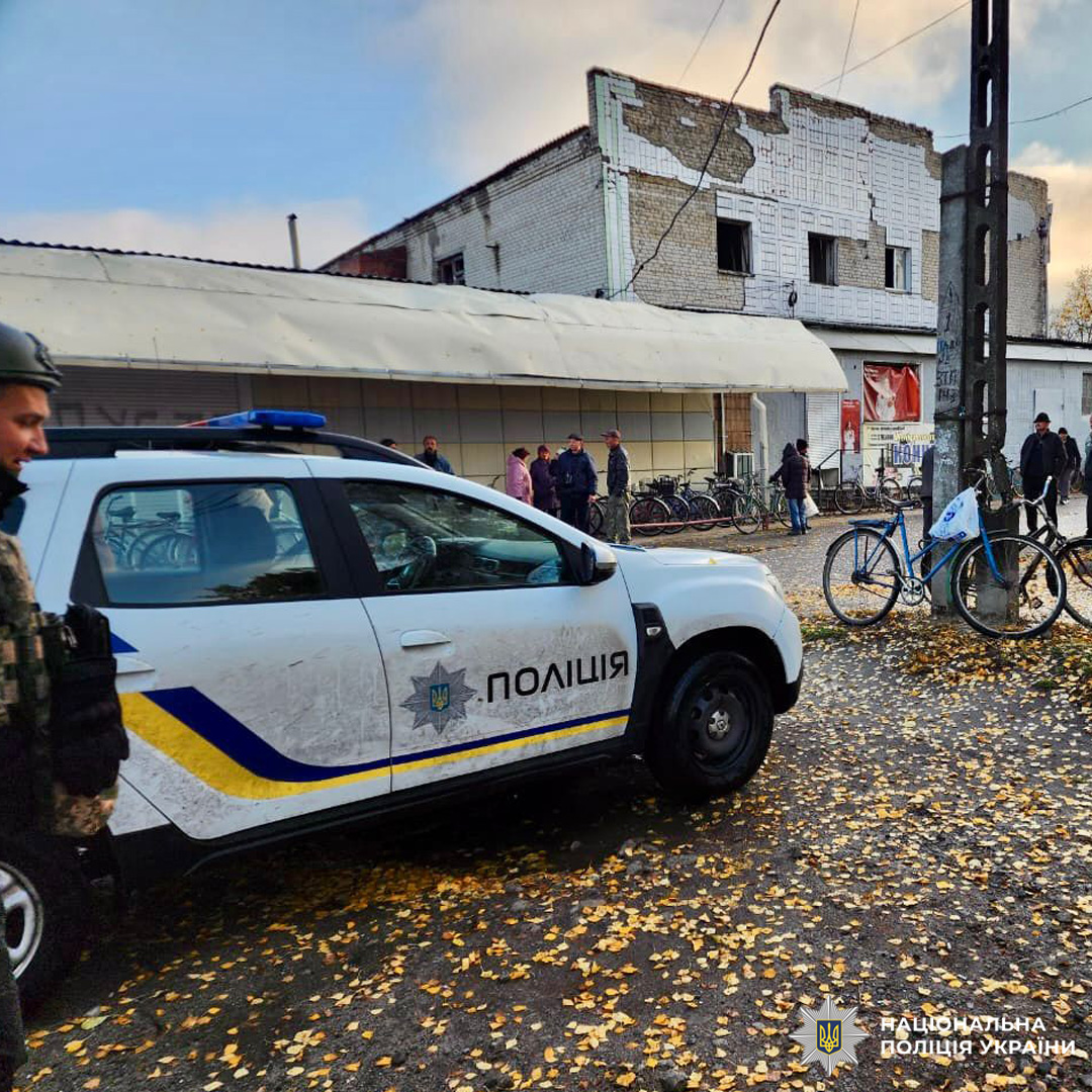 Поліцейське авто на вулиці, поруч поліцейський, люди біля будівлі, осіннє листя на землі.