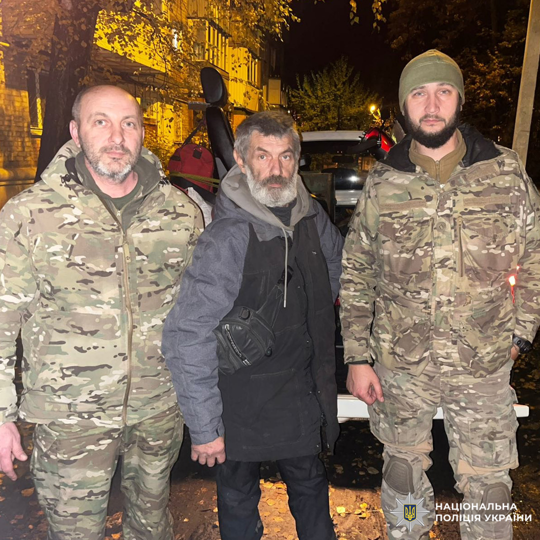 Співробітники Національної поліції України в камуфльованій формі разом із цивільним чоловіком у темному одязі. Група сфотографована вночі на тлі автівки та осінніх дерев.