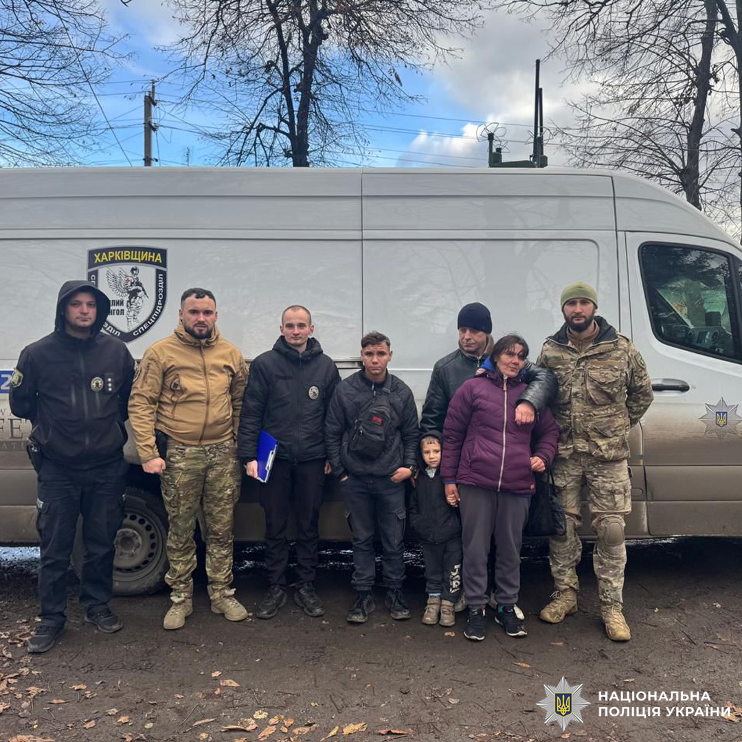 Співробітники Національної поліції України на Харківщині разом із евакуйованими цивільними чоловіком, жінкою, підлітками та дитиною. Група стоїть біля службового мікроавтобуса з емблемою поліції.