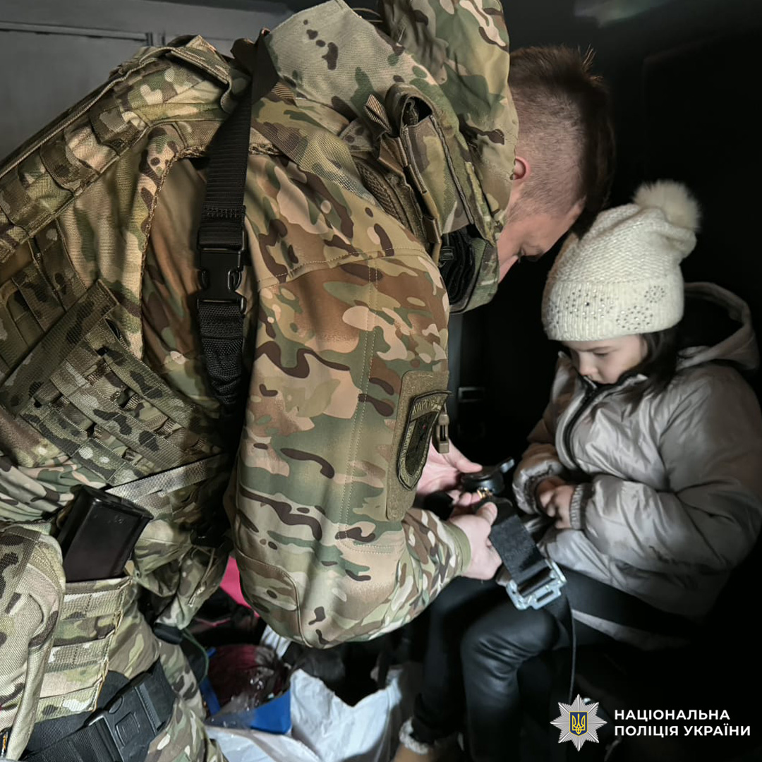 Співробітник Національної поліції України в тактичному спорядженні допомагає дитині в зимовому одязі всередині службового транспорту. На тлі сумки та речі, що свідчать про евакуацію.