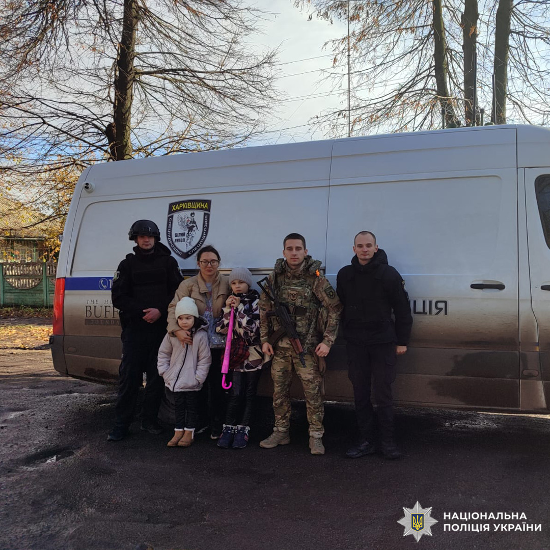 Співробітники Національної поліції України на Харківщині разом із цивільними жінкою та двома дітьми біля службового мікроавтобуса з емблемою поліції. Один із дітей тримає рожеву парасольку.