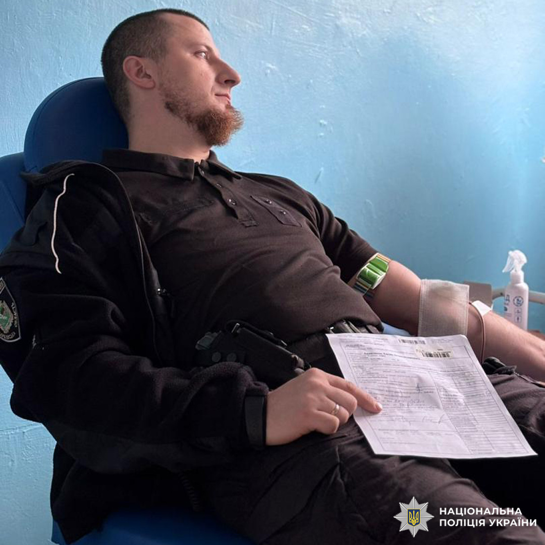 Співробітник поліції здає кров у медичному закладі. Донор перебуває у спеціальному кріслі, тримає документ, пов’язаний із процедурою. До руки під’єднано систему для забору крові. Поруч антисептичні засоби та медичне обладнання.