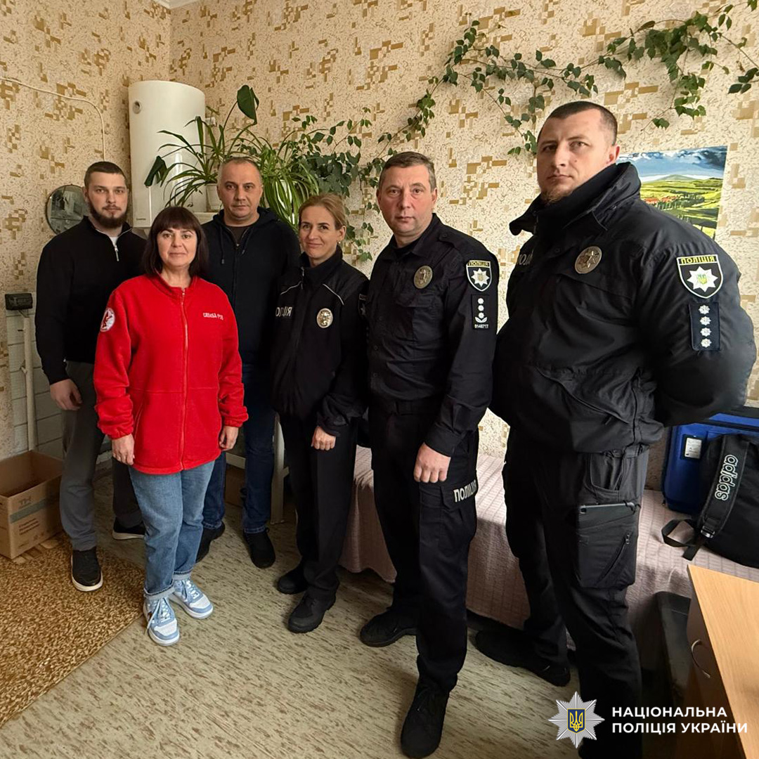 Правоохоронці у службовій формі здійснюють візит до приміщення з цивільними особами. У кімнаті ліжко, коробка, сумка та кімнатні рослини. На стіні декоративне панно.