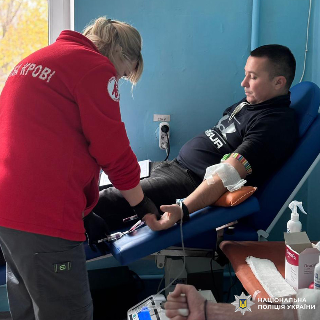 Поліцейський здає кров у медичному закладі. Медичний працівник Служби крові здійснює забір. На передньому плані медичне обладнання та засоби дезінфекції.