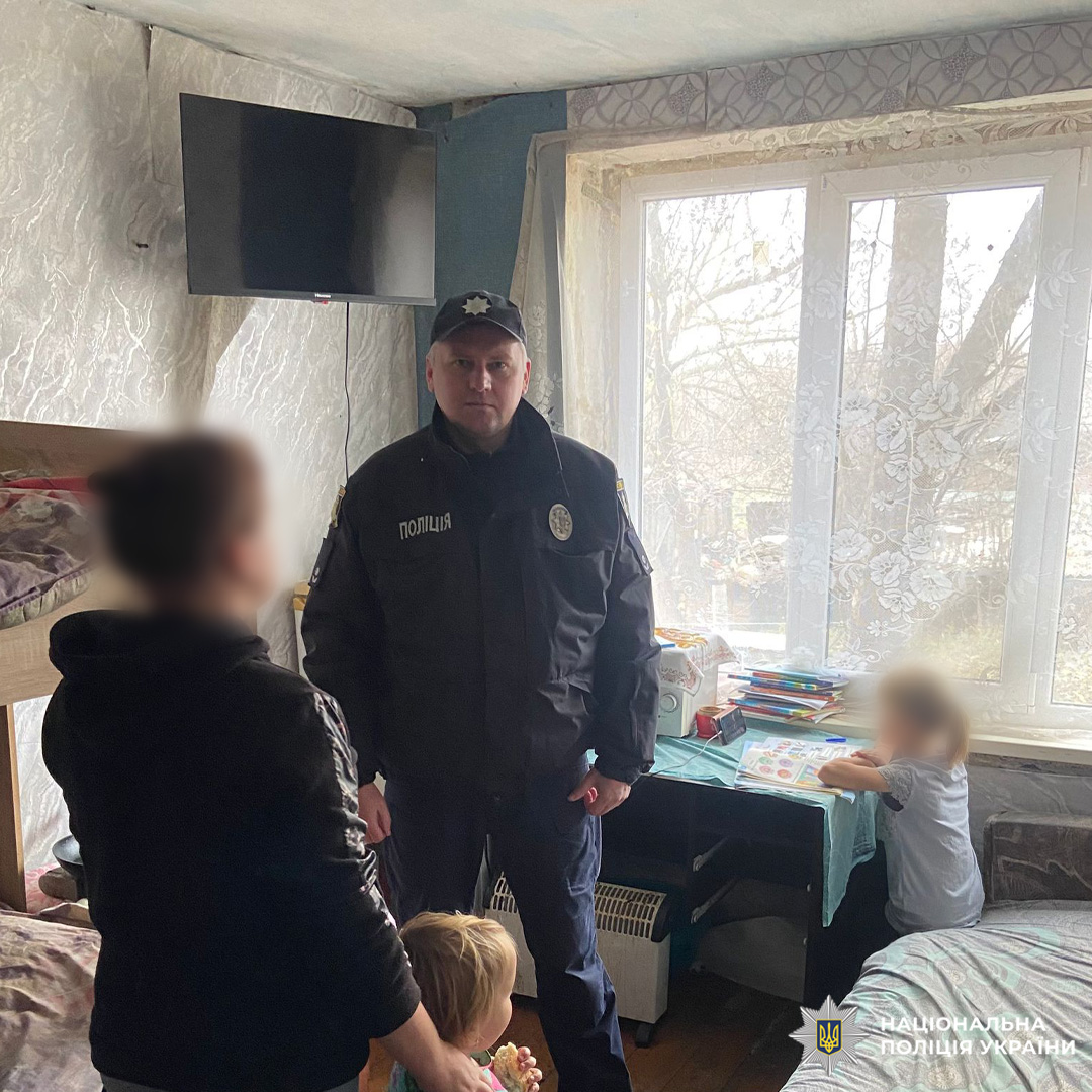Співробітник Національної поліції України проводить превентивну роботу з багатодітною родиною у житловому приміщенні. На фото троє дітей у кімнаті з двоярусним ліжком, телевізором та навчальним столом.