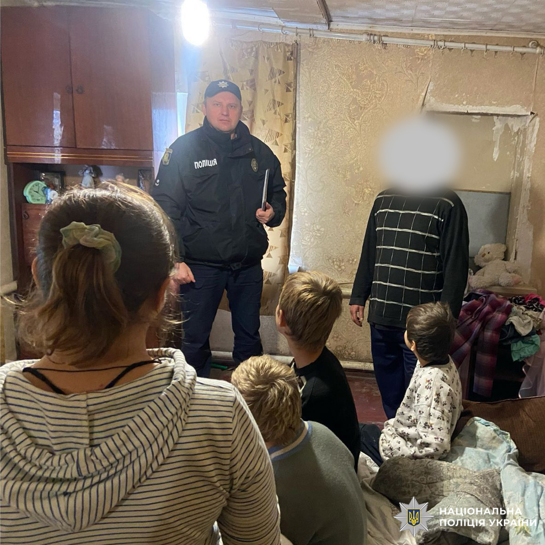 Співробітник Національної поліції України проводить превентивну бесіду з родиною у житловому приміщенні. На фото діти та дорослий із прихованим обличчям у кімнаті з побутовими речами.