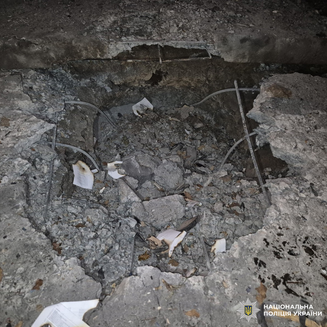 Велика вирва у бетонному покритті, ймовірно спричинена вибухом. Навколо уламки, арматура та розкидані папери.