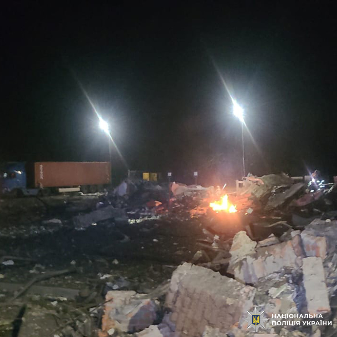 Нічна сцена руйнування: серед уламків горить вогонь, освітлюючи територію. На фоні вантажівка, контейнер і вуличні ліхтарі.