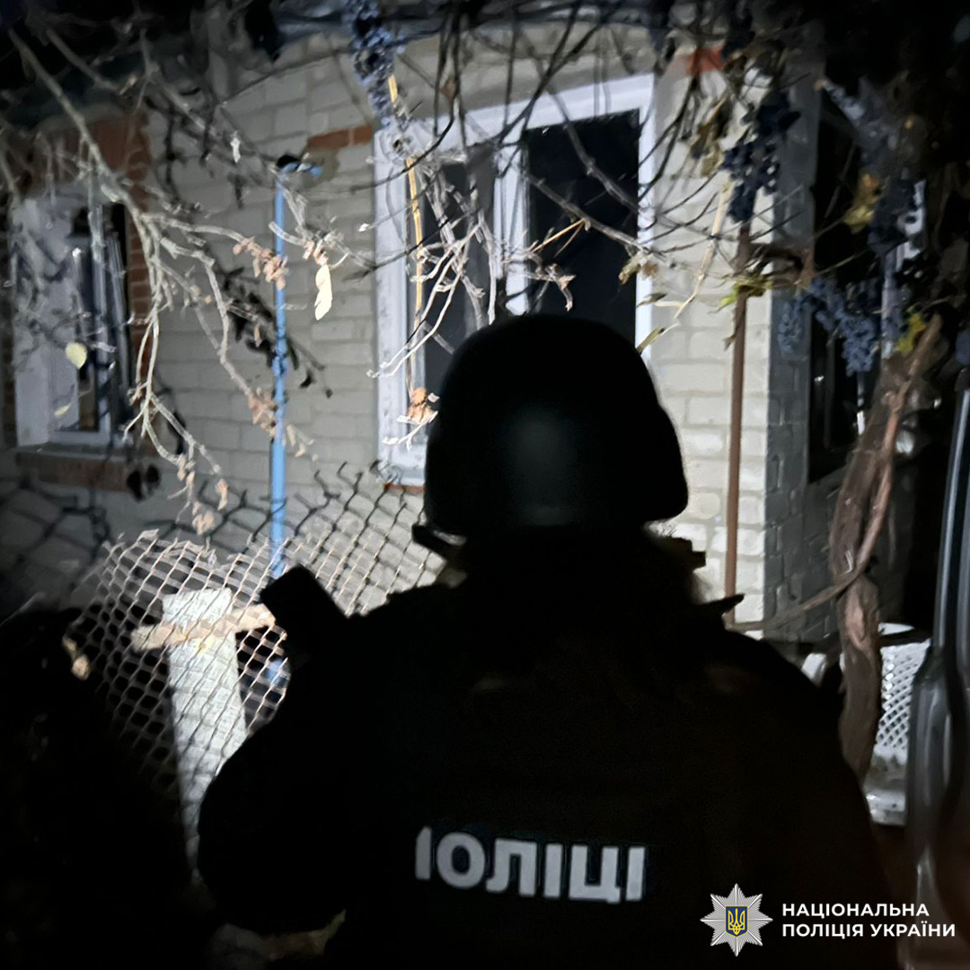 Поліцейський у шоломі та бронежилеті стоїть біля пошкодженого вікна цегляної будівлі. На передньому плані зігнутий металевий паркан, уламки та гілки з сухим листям.