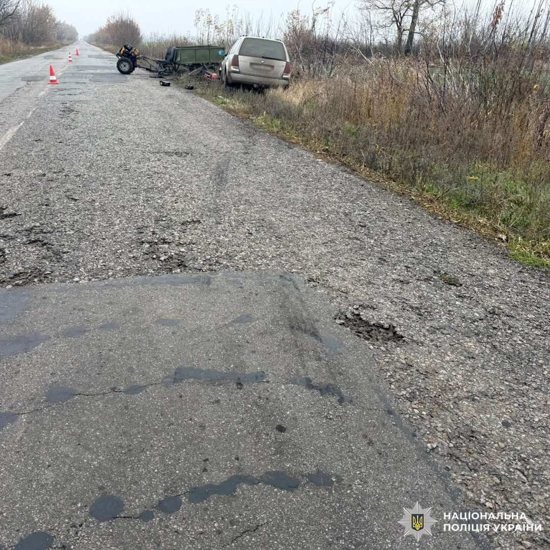 Сцена дорожньо-транспортної пригоди на сільській дорозі: пошкоджений мотоцикл, причіп і легковий автомобіль на узбіччі. Асфальт нерівний і вологий, навколо суха рослинність.