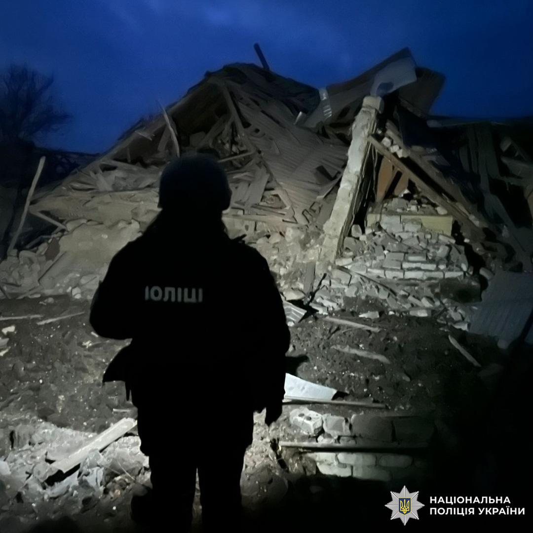 Поліцейський у захисному шоломі оглядає місце нічної руйнації будівлі. На фоні уламки дерев’яних конструкцій і цегли, освітлені прожектором.