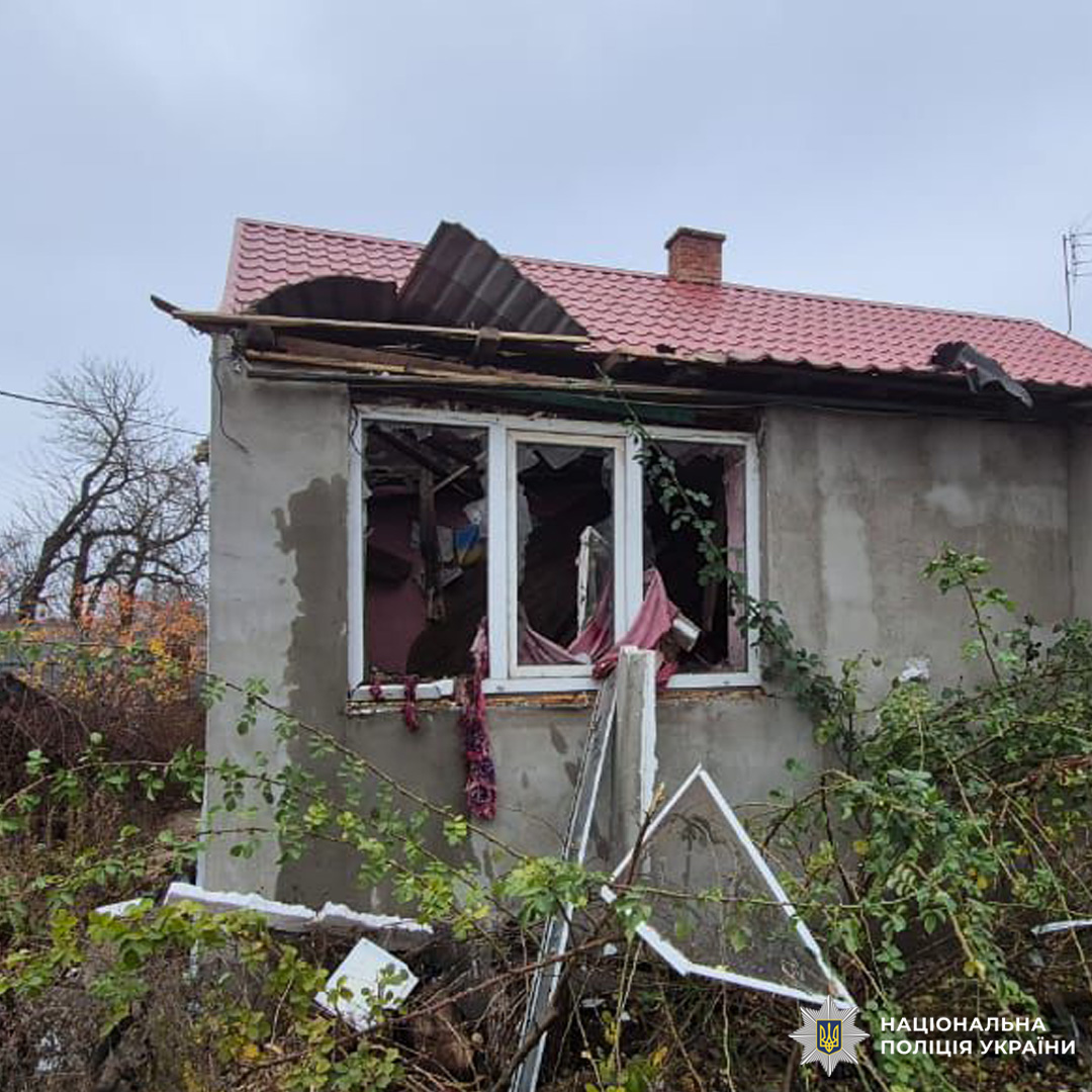Пошкоджений житловий будинок із черепичним дахом: верхня частина даху зруйнована, вікна вибиті, крізь скло звисають штори. Перед фасадом уламки конструкцій і рослинність.