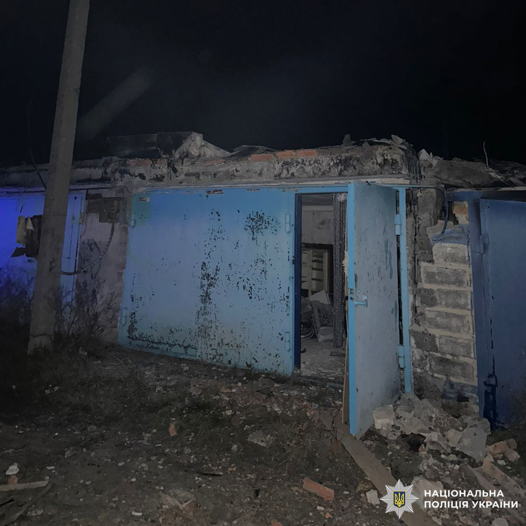 Нічна сцена пошкодженої будівлі з частково обваленим дахом і розкиданими уламками. Сині металеві двері, одна з яких відчинена, відкривають доступ до приміщення з видимими меблями. Територія освітлена штучним світлом.