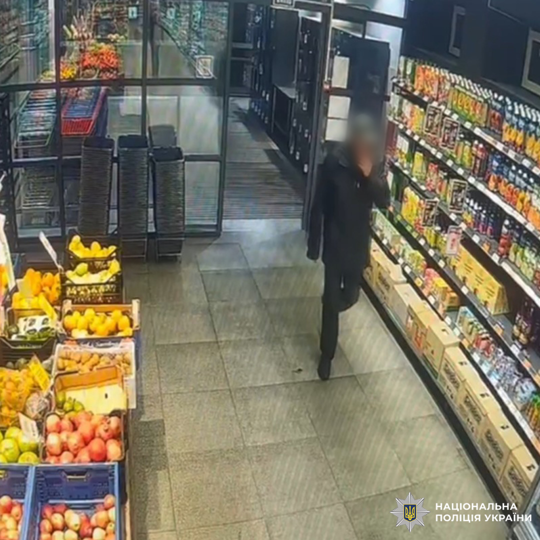 Кадр із камери спостереження в продуктовому магазині: особа в темному одязі рухається між стелажами з товарами. Ліворуч ящики з фруктами.