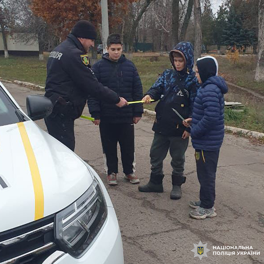 Поліцейський на вулиці передає дітям світловідбивні стрічки. Діти в зимовому одязі уважно слухають. На фоні службовий автомобіль і дерева.