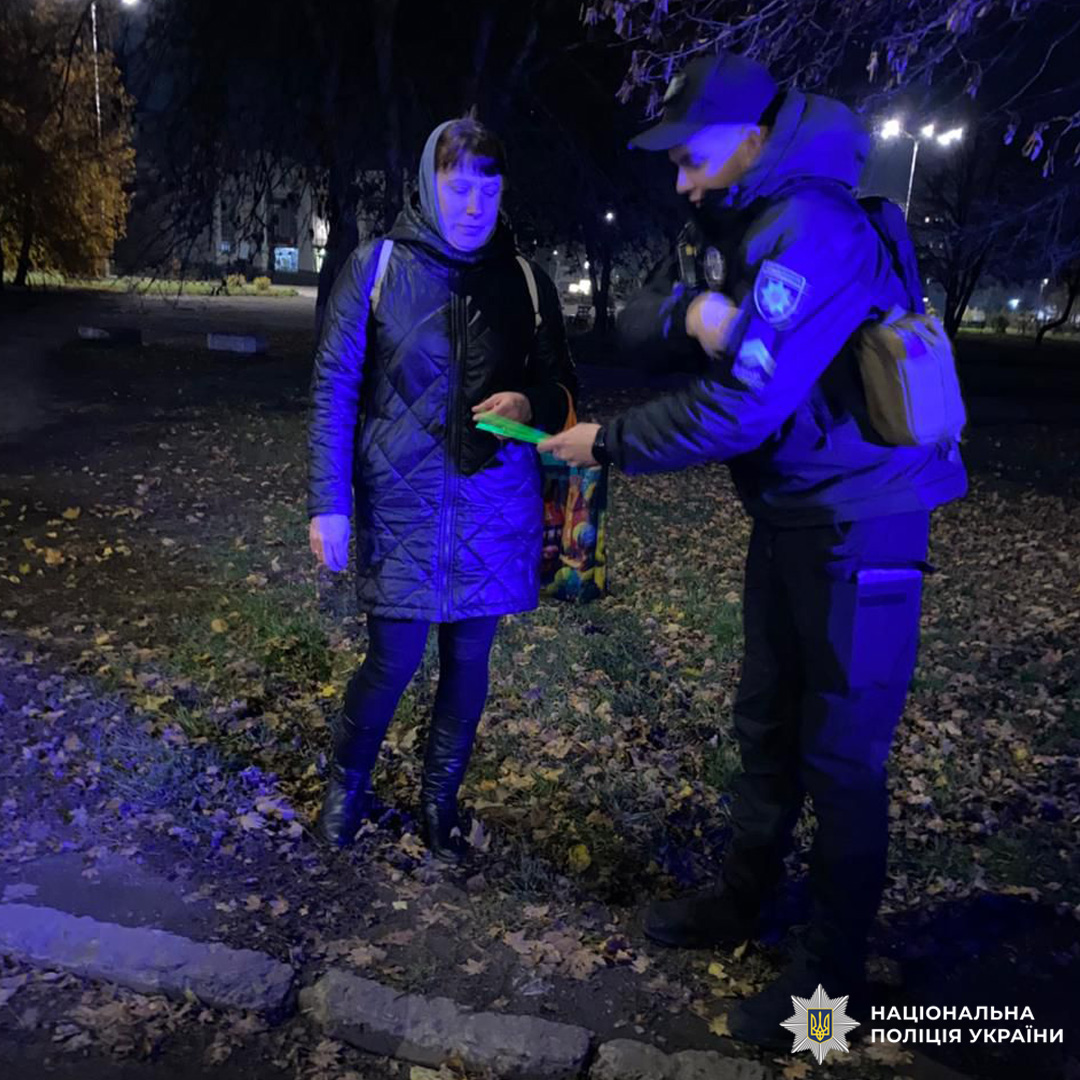 Особа в темному пальті з кольоровою сумкою спілкується з поліцейським. На землі опале листя, на фоні дерева та лавки.