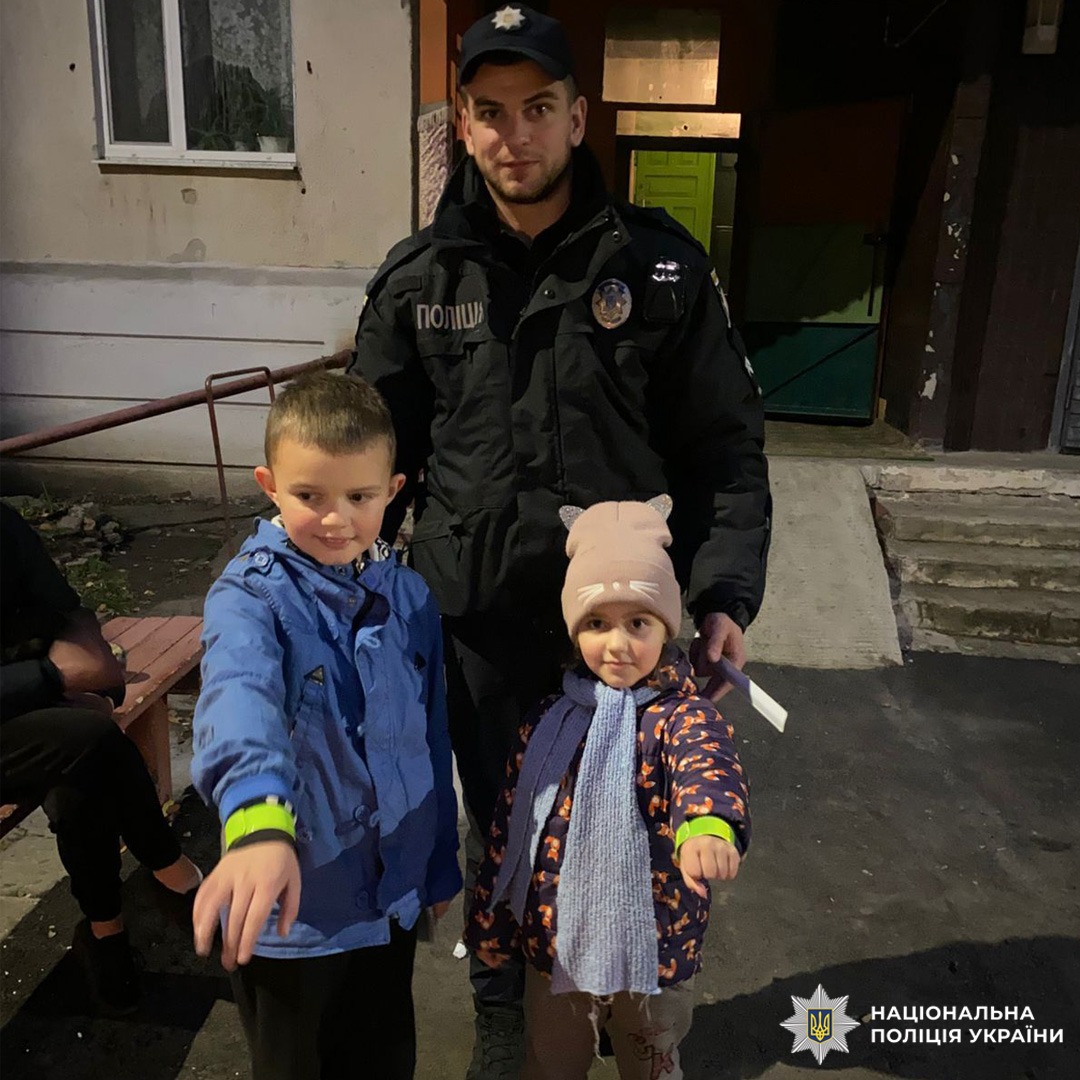 Поліцейський у формі поруч із двома дітьми біля житлової будівлі. На фоні лавка, сходи та вхідні двері.