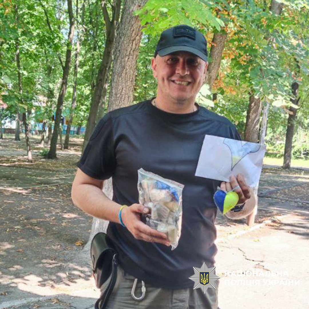 У парку на тлі дерев та асфальтованої доріжки стоїть працівник поліції. Він тримає прозорий пакет із дрібними предметами, аркуш із малюнком та дві кульки жовту і синю.