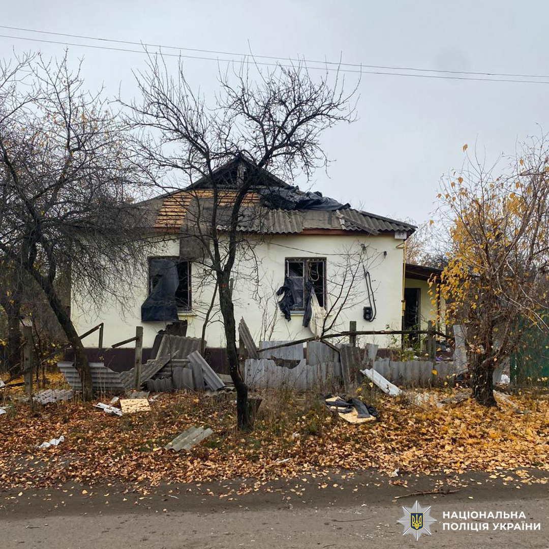 Пошкоджений житловий будинок із частково зруйнованим дахом і вибитими вікнами. Видимі сліди пожежі та задимлення, навколо уламки паркану та будівельні матеріали. Осіннє листя вкриває землю, дерева без листя.