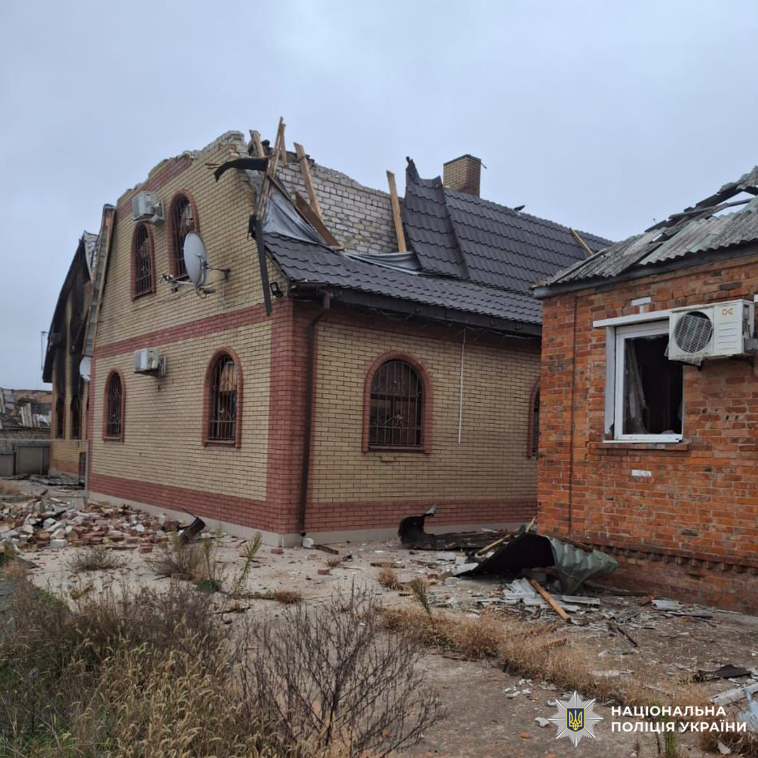 Житловий будинок із серйозними пошкодженнями, частково обвалений дах, зруйновані вікна, уламки на подвір’ї. Будівля цегляна, з арочними вікнами та супутниковими антенами на фасаді. Поруч ще одна пошкоджена споруда.
