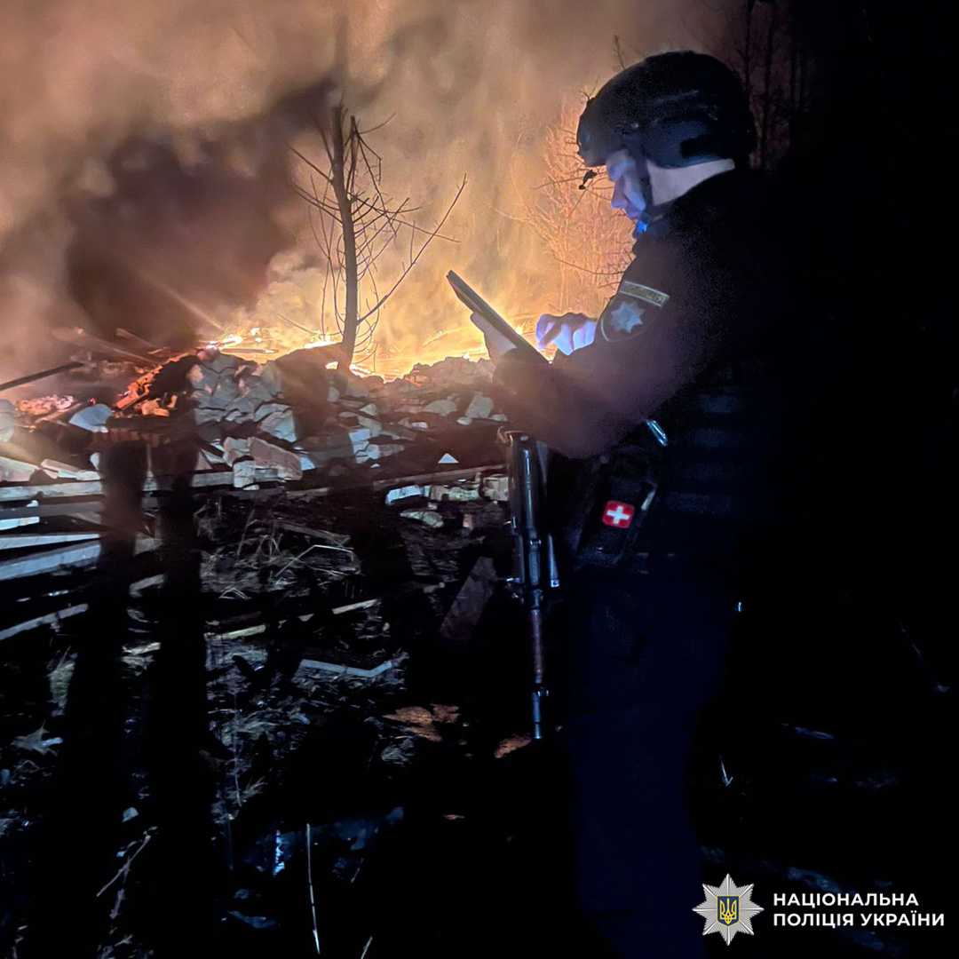 Співробітник Національної поліції України в тактичному спорядженні стоїть перед палаючими уламками вночі. На бронежилеті медична підсумка з червоним хрестом, в руках вогнепальна зброя та пристрій зв’язку. Потужне полум’я охоплює дерев’яні конструкції та цеглу, густий дим здіймається вгору.