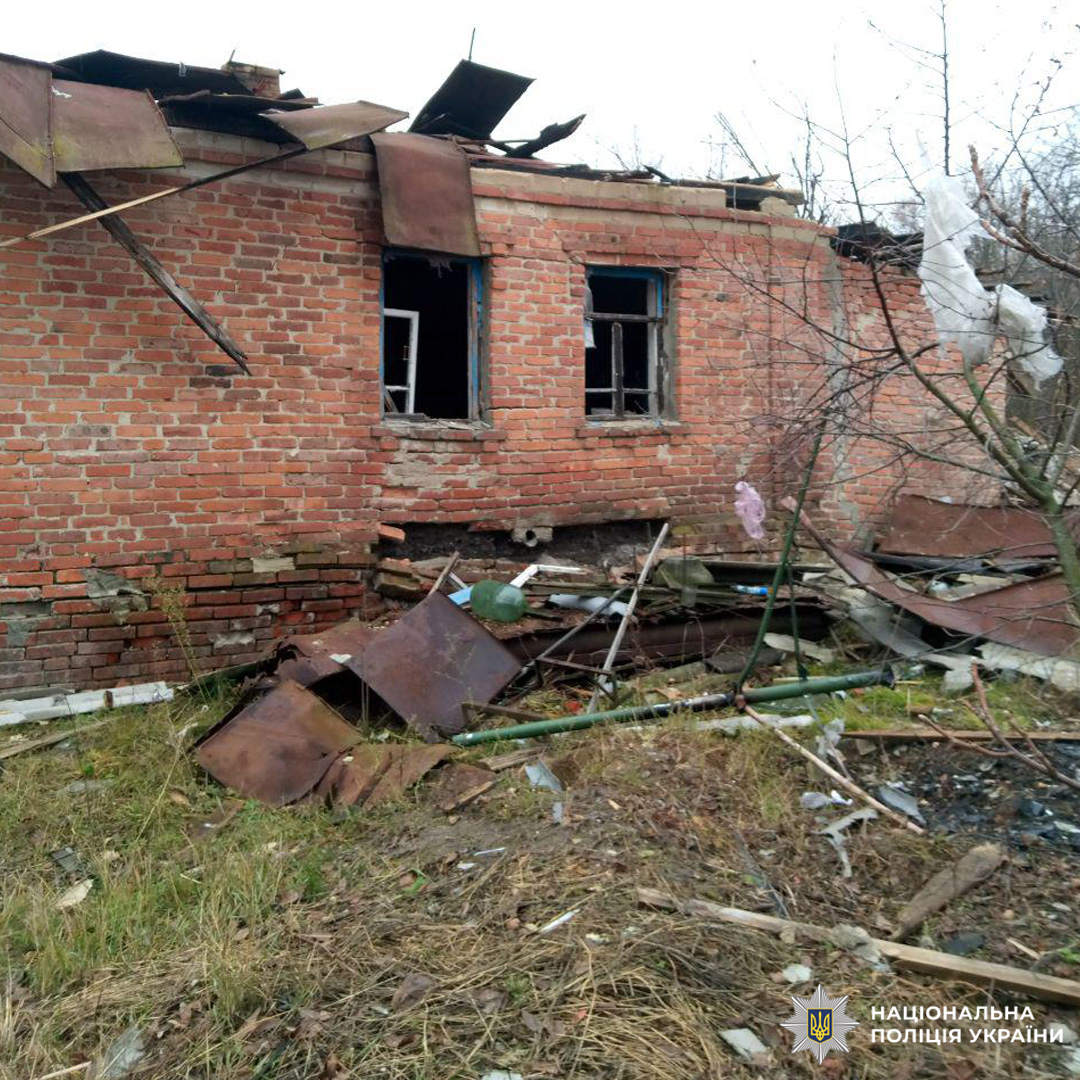 Цегляна будівля з частково обваленим дахом, зруйнованими вікнами та розкиданими уламками. Стіни мають ознаки руйнування, територія навколо заросла травою, поруч голі дерева.