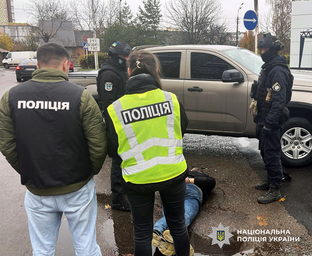 на асфальті лежить затриманий фігурант, поруч стоять поліцейські та автомобіль