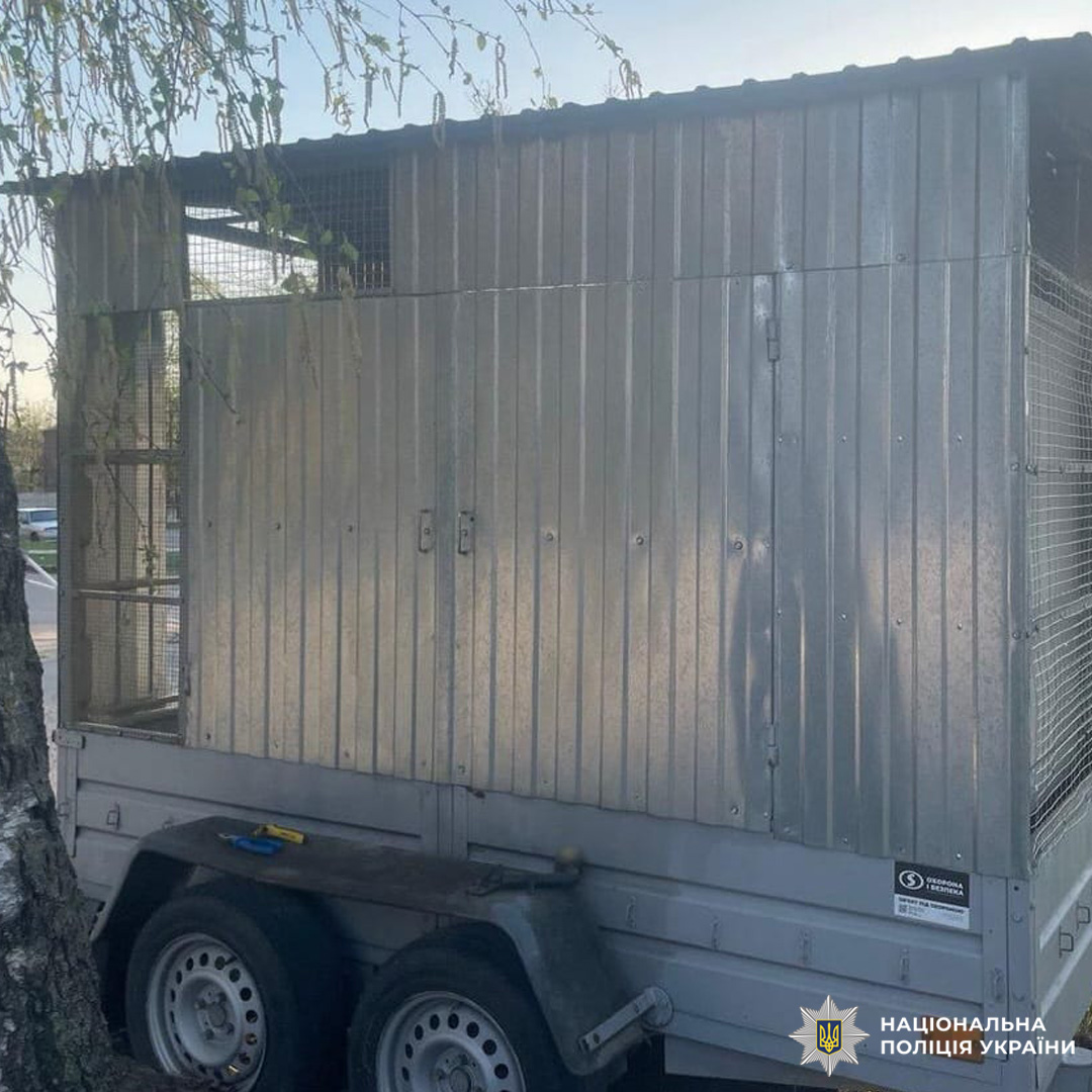 Металевий причіп із вентиляційною решіткою, зафіксований Національною поліцією України. Причіп має подвійні осі, бічні двері та ознаки використання для транспортування спецвантажів.