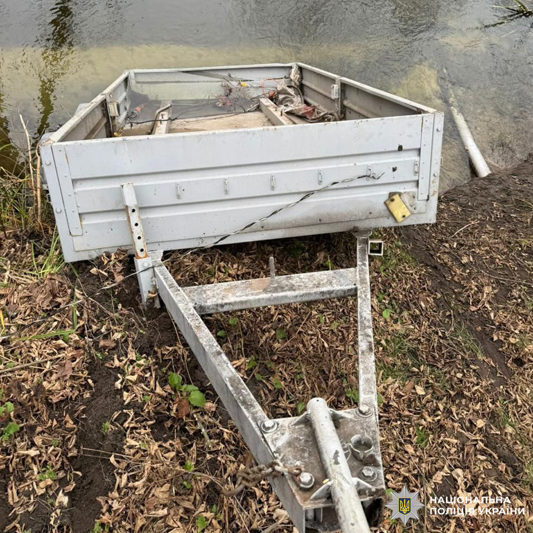 Пошкоджений причіп на березі водойми, зафіксований співробітниками Національної поліції України. Видимі деформації конструкції, задня частина спрямована до води.