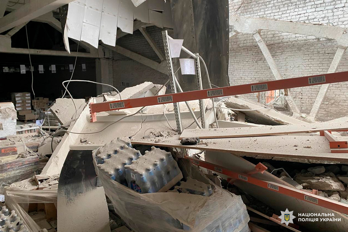 На фото внутрішнє приміщення складу з ознаками серйозних пошкоджень. Частково обвалена стеля, пошкоджені стелажі, на підлозі уламки конструкцій та палети з бутильованою водою. Видимі маркування на стелажах.