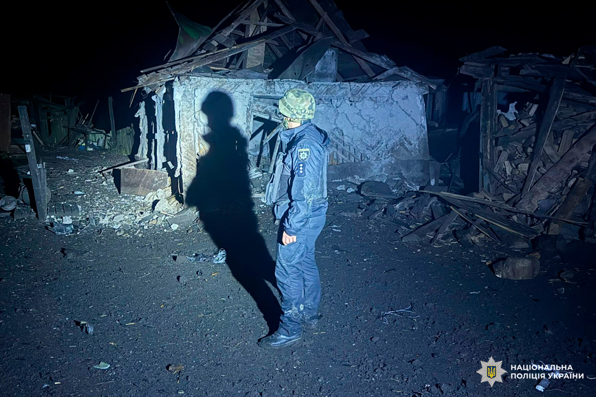 На фото співробітник Національної поліції України в захисному спорядженні, який перебуває біля зруйнованої будівлі. Зйомка здійснена вночі. Видимі уламки дерев’яних конструкцій та серйозні пошкодження споруди.