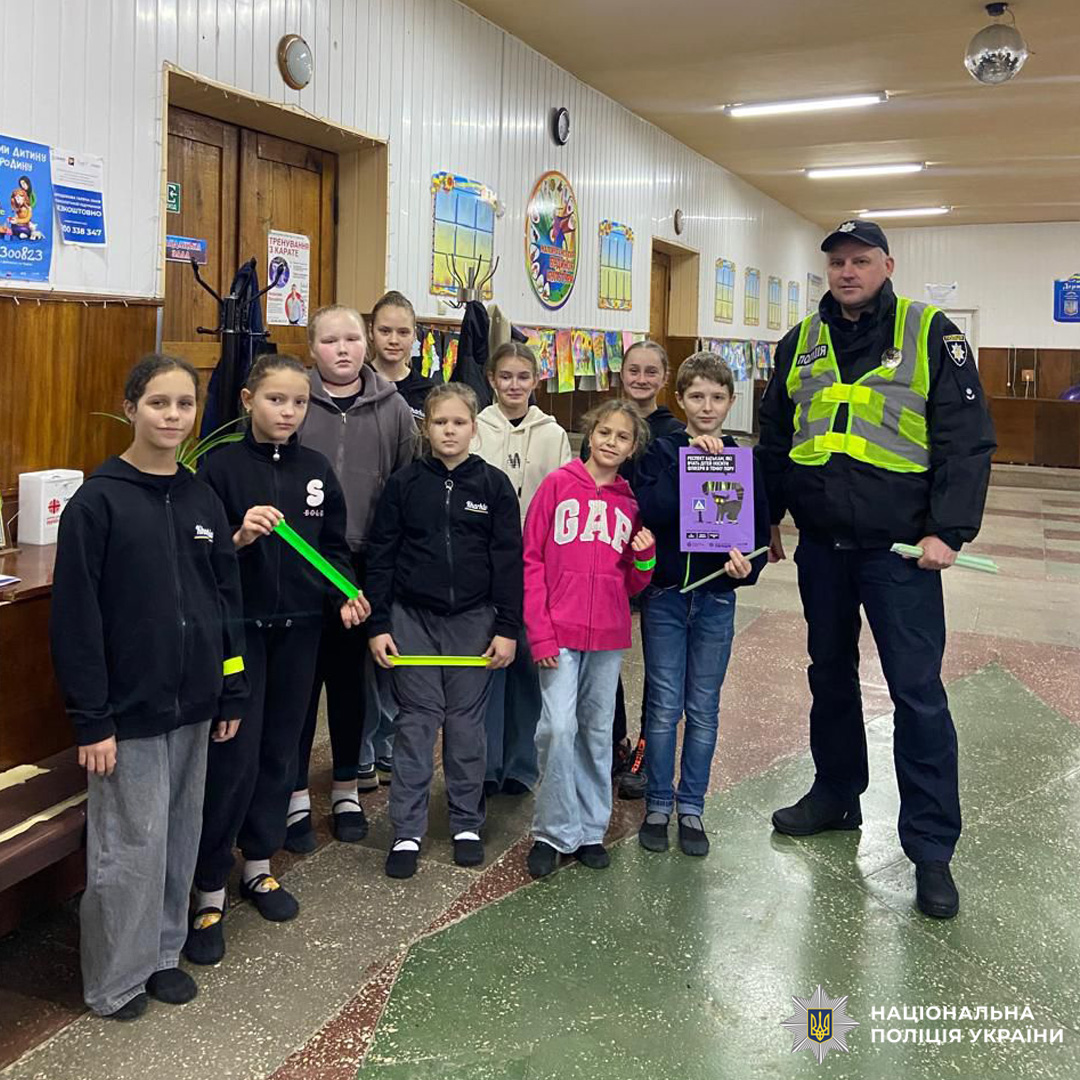 Група дітей у приміщенні з білими панелями та яскравими декораціями разом із поліцейським, дитина тримає зелений світловідбивний елемент. Захід організовано Нацполіцією України для навчання з безпеки.