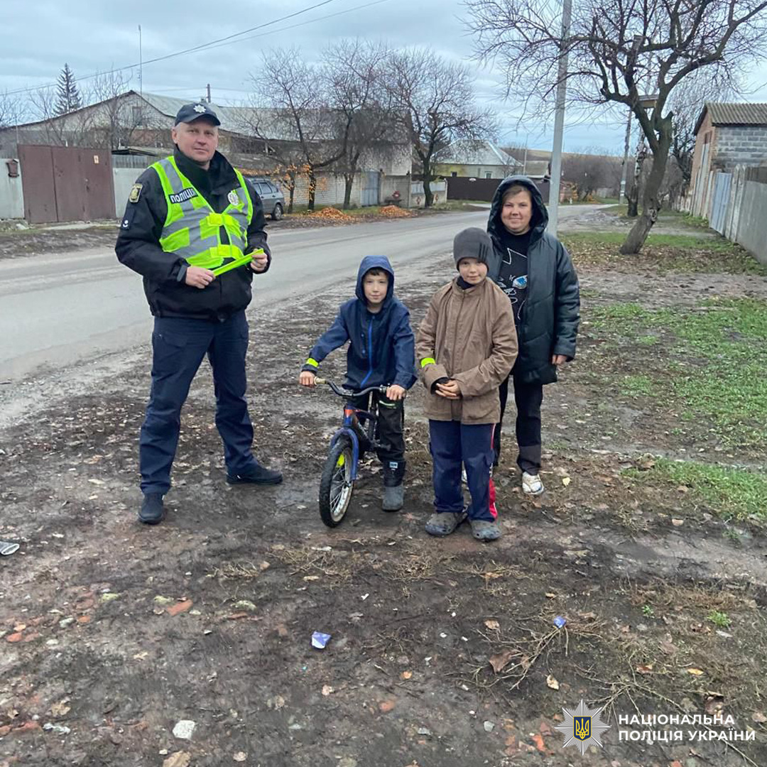 Поліцейський у світловідбивному жилеті спілкується з трьома дітьми на узбіччі дороги в житловому районі. Один із дітей на велосипеді, всі одягнені по-зимовому, на фоні будинки та голі дерева