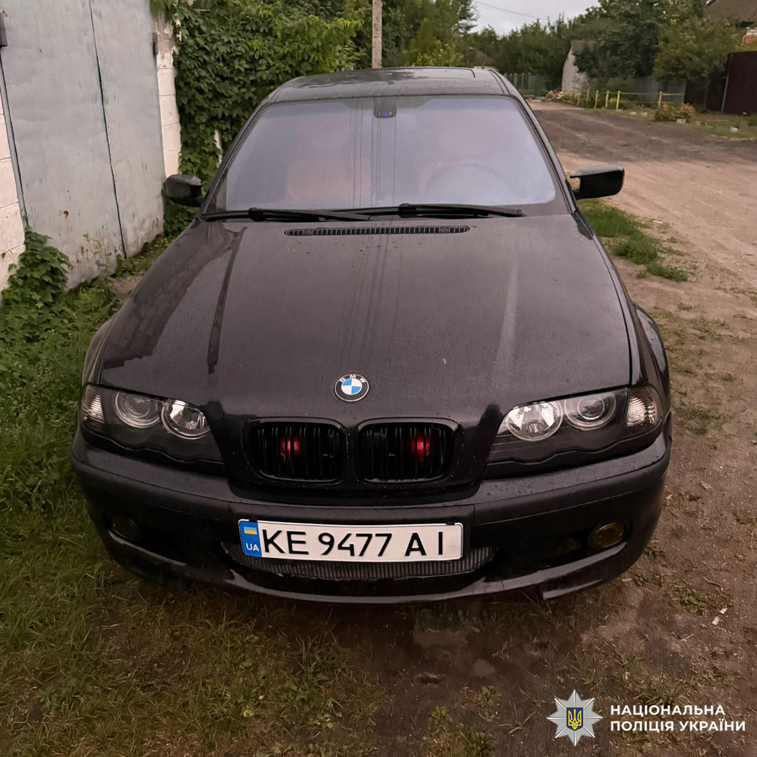 Чорний автомобіль BMW з українськими номерними знаками "KE 9477 АІ" припаркований на траві біля ґрунтової дороги.Авто мокре, ймовірно, після дощу.