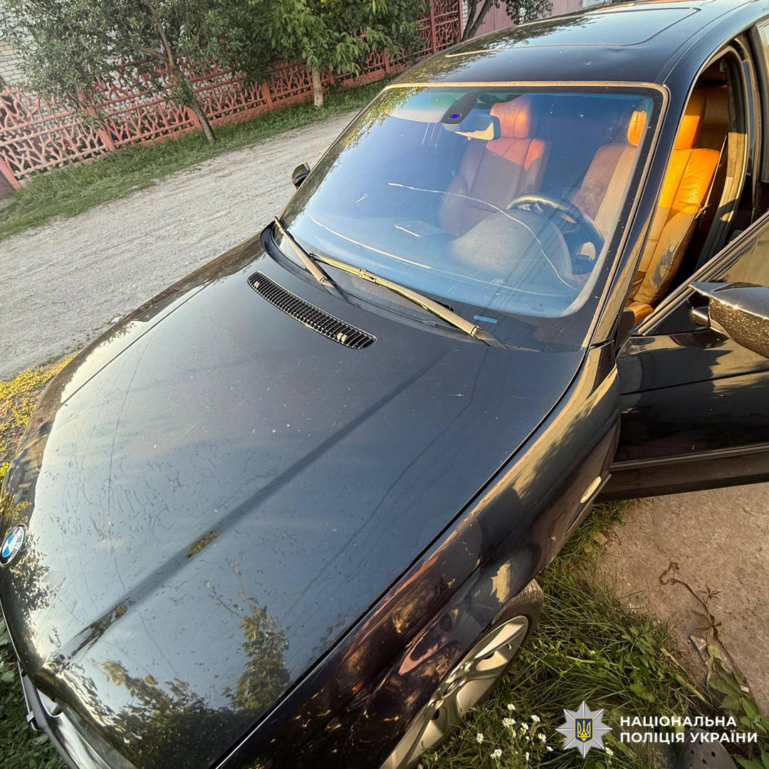 Чорний автомобіль BMW з українськими номерними знаками "KE 9477 АІ" припаркований на траві біля ґрунтової дороги.Авто мокре, ймовірно, після дощу.