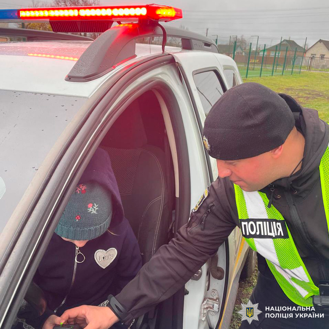 Співробітник Національної поліції України у формі та світловідбивальному жилеті, який взаємодіє з дитиною в службовому автомобілі. Дитина одягнена в зимову шапку та куртку з нашивкою "LOVE".