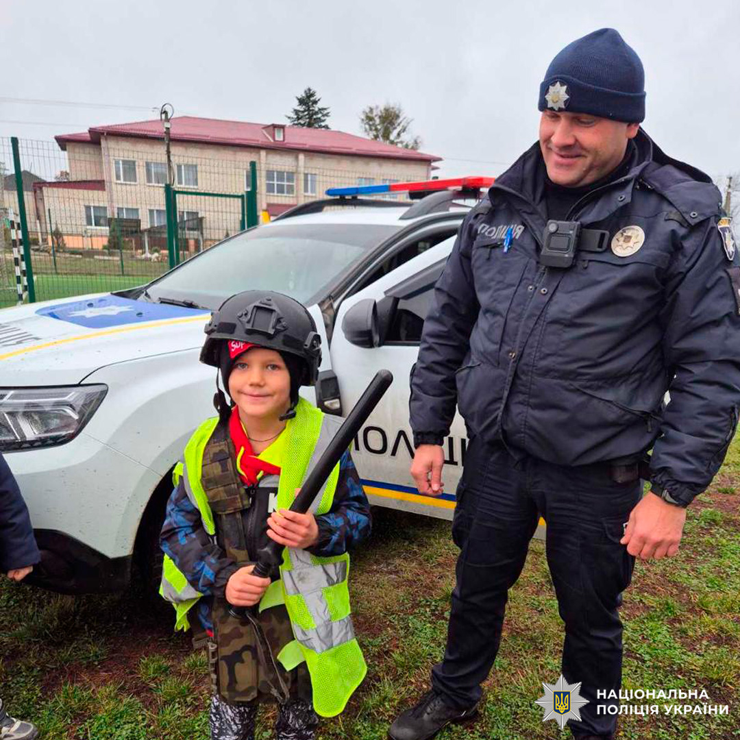 Дитина в поліцейському шоломі, світловідбивальному жилеті та з кийком, яка стоїть біля співробітника Національної поліції України. На задньому плані службовий автомобіль, огорожа та будівлі.