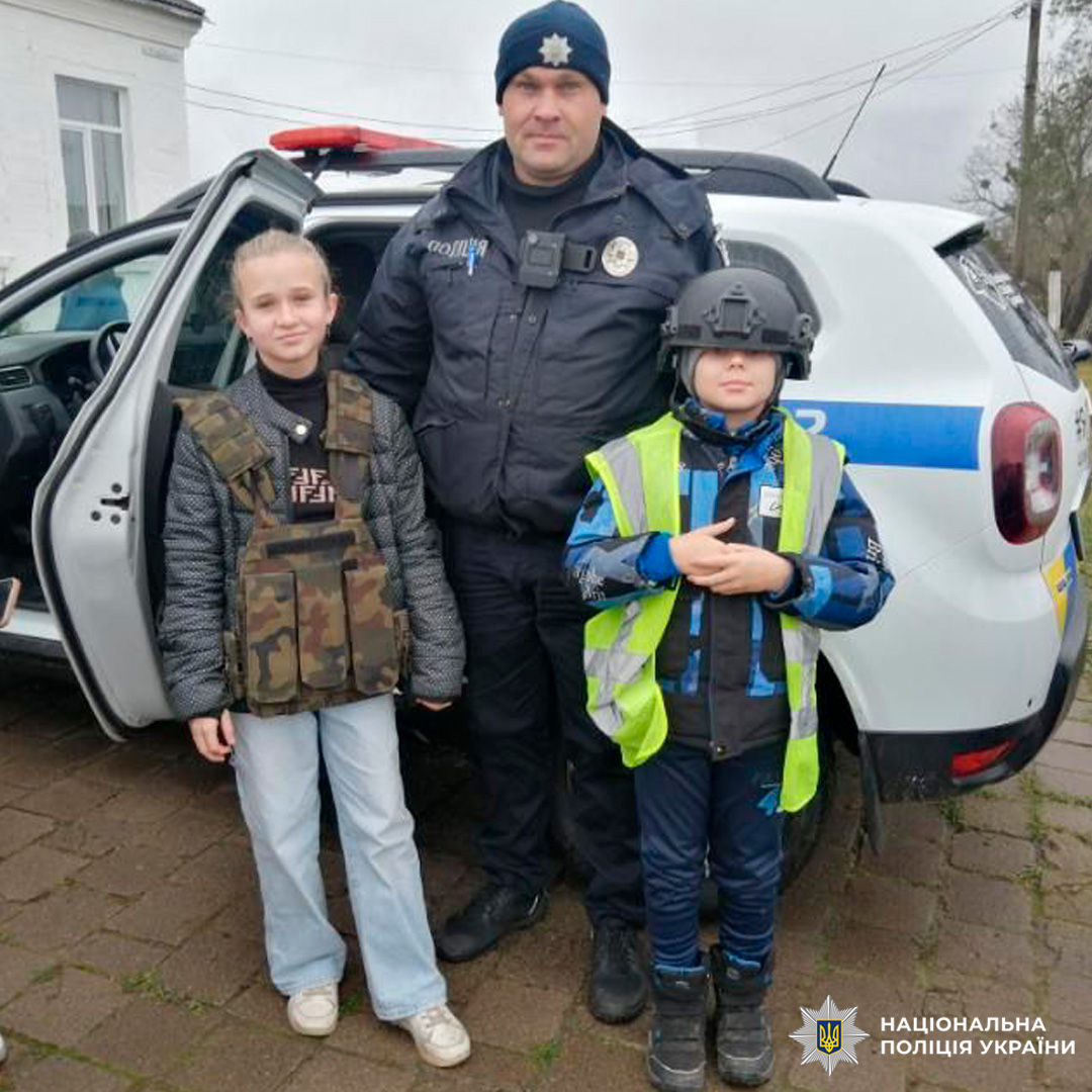 Поліцейський разом із двома дітьми в екіпіруванні позують біля службового авто.