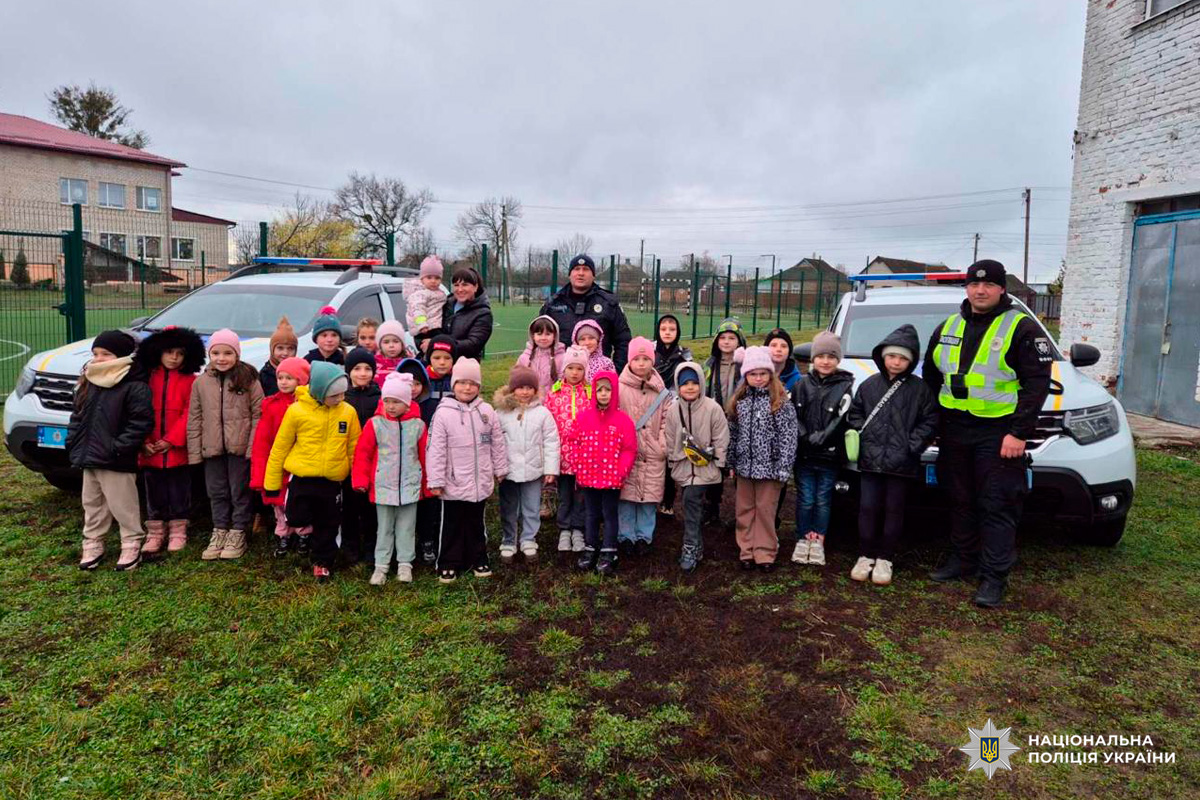 Група дітей і дорослих, серед яких співробітники Національної поліції України у формі та світловідбивальних жилетах. Усі стоять перед двома службовими автомобілями.