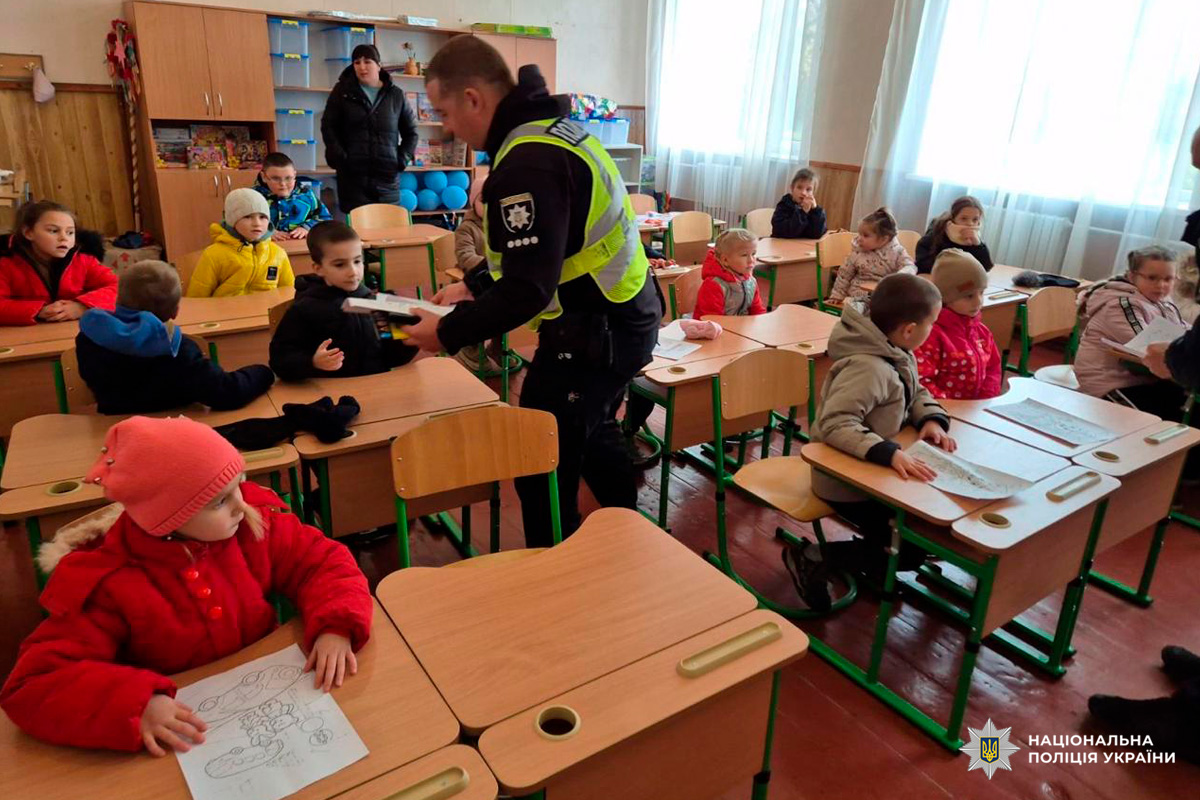 Співробітник Національної поліції України, який взаємодіє з дітьми в класі, роздаючи друковані матеріали. Учні одягнені в теплий одяг, займаються розмальовками. У класі навчальні матеріали, іграшки та шафи для зберігання.