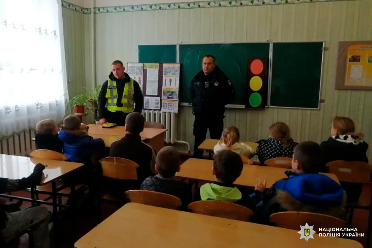 Двоє співробітників Національної поліції України, які проводять заняття для учнів у класі. На дошці схема світлофора та навчальні плакати. Діти сидять за партами, уважно слухаючи.