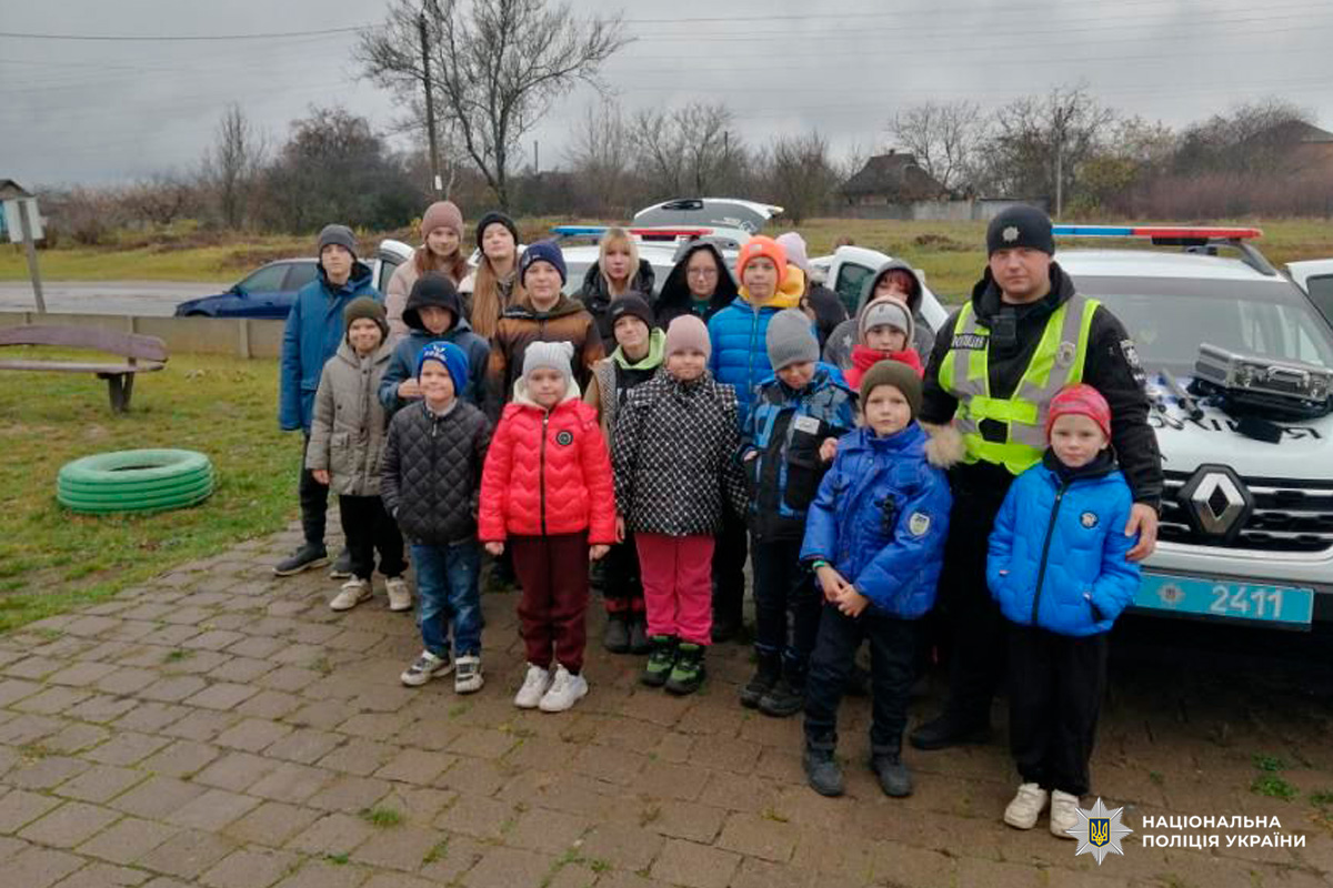 Група дітей і дорослих, серед яких співробітники Національної поліції України у формі та світловідбивальних жилетах. Усі стоять перед службовим автомобілем на відкритій території.