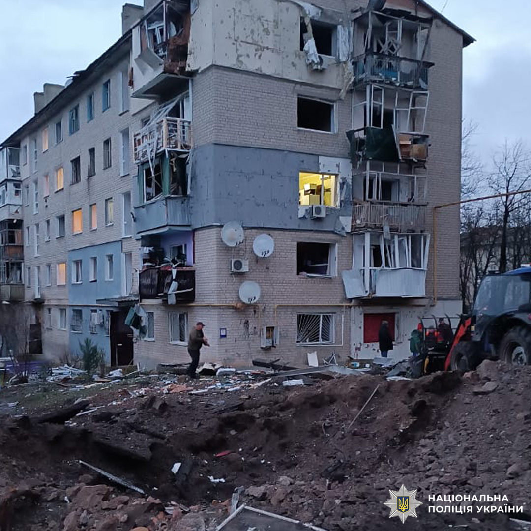 Багатоповерховий житловий будинок із серйозними пошкодженнями фасаду, вікон і балконів, частково зруйнований внаслідок потужного вибуху. На передньому плані велика вирва, уламки та кілька людей, зокрема представники екстрених служб.