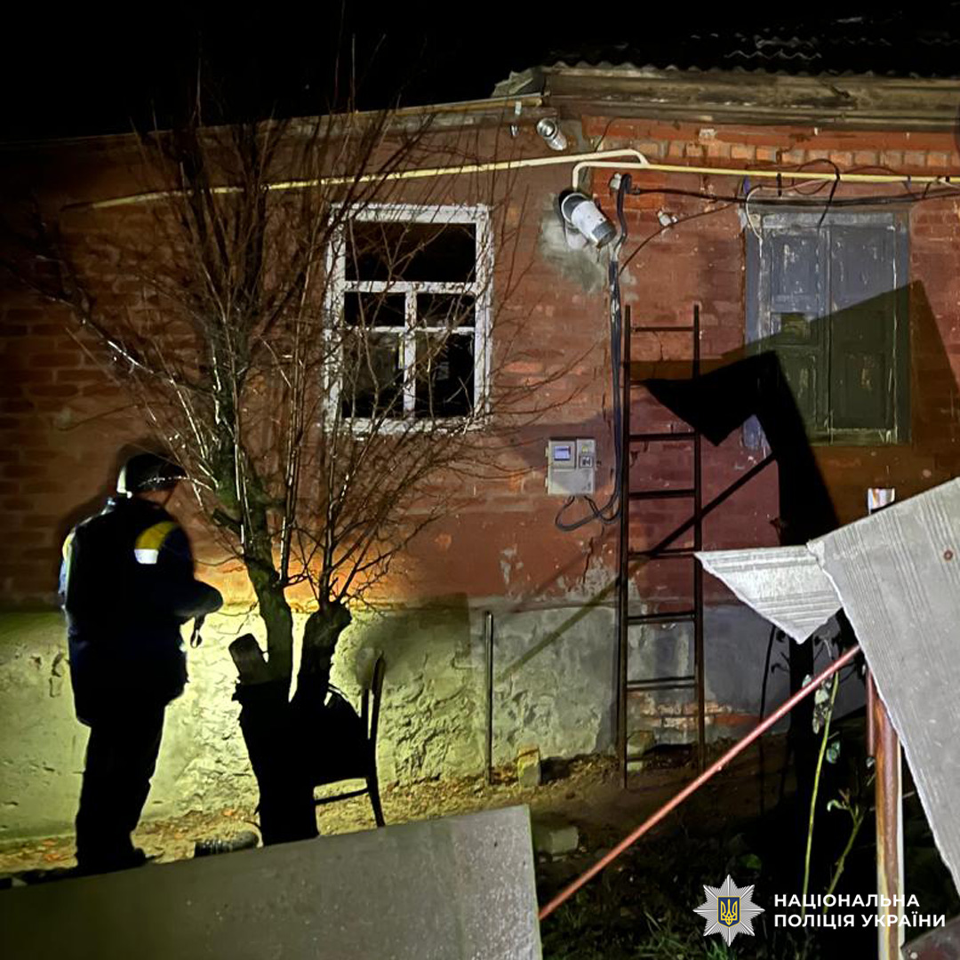 Поліцейський, освітлює пошкоджену цегляну будівлю ліхтарем. Видно розбите вікно, зламані двері та драбину, що спирається на стіну. На землі металеві листи, кабелі та інші предмети.