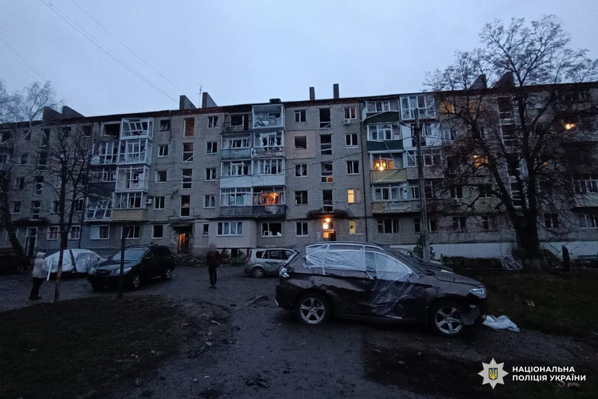 Житловий багатоповерховий будинок із пошкодженими вікнами та балконами, перед яким стоять автомобілі, частково накриті плівкою. На землі уламки, одна людина проходить повз.