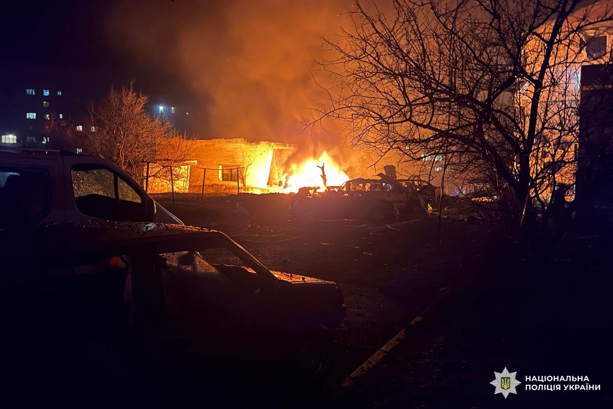 Кілька пошкоджених автомобілів, один із них охоплений вогнем. Яскраве полум’я освітлює навколишню територію, видно будівлі та голі дерева. Один із будинків охоплений вогнем.