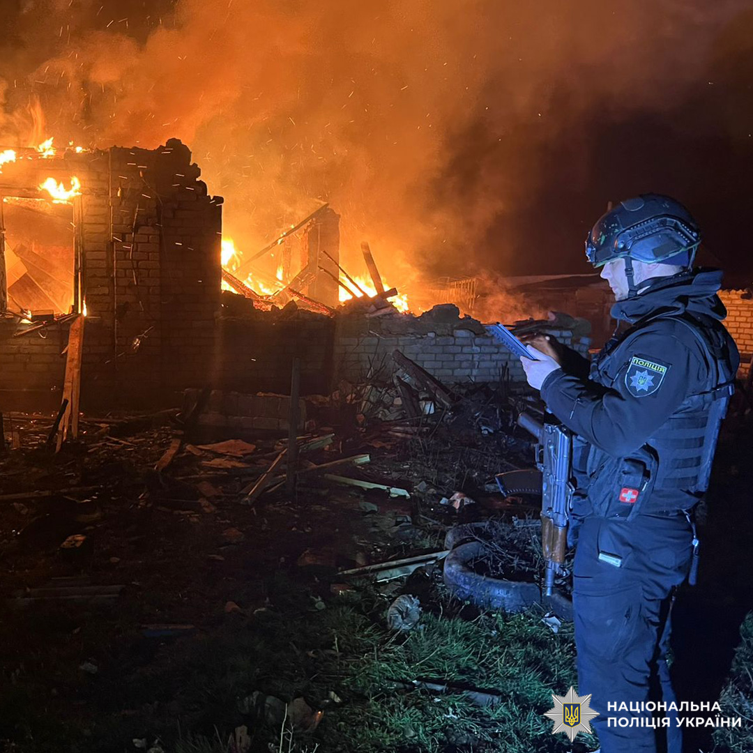 Палає будівля з цегли та дерева. Вогонь охопив дах і стіни, частина конструкцій зруйнована. На передньому плані озброєний співробітник у тактичному спорядженні, який дивиться на пристрій у руках. Навколо уламки, густий дим і яскраве світло від пожежі.