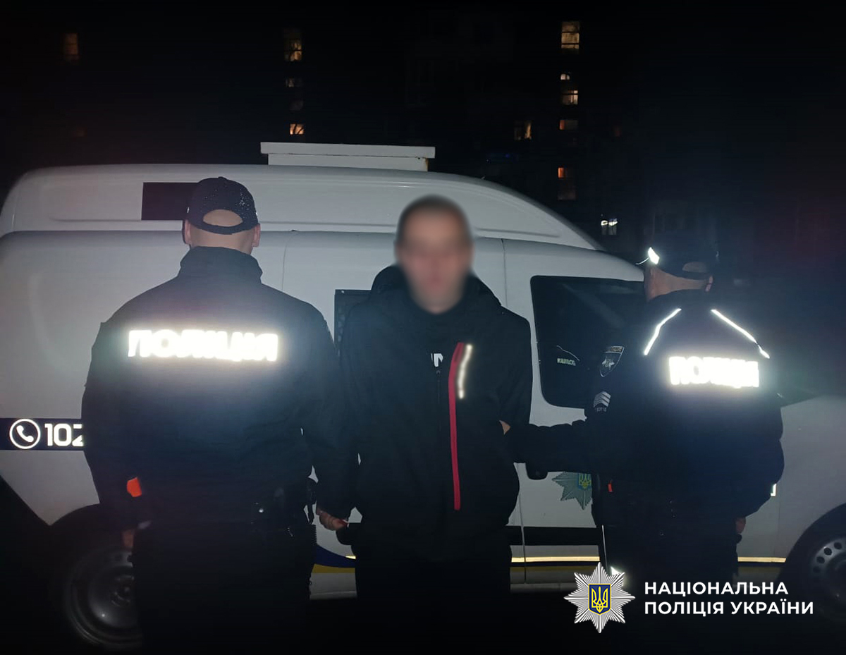 У темну пору доби двоє поліцейських стоять біля службового автомобіля та тримають затриманого чоловіка.